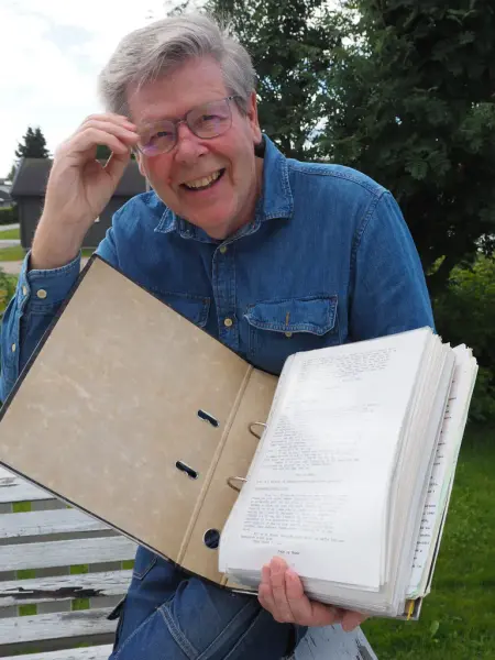 En mann med grått hår, briller og dongeriskjorte holder en åpen perm fylt med mange ark. Trær og hagebenk i bakgrunn.