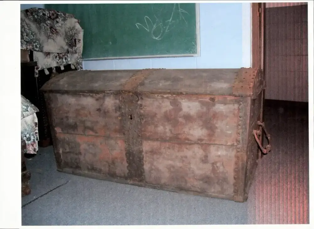 En stor kiste med buet lokk,  jernbeslag og en rester av rødaktig maling står på et gulv. Sammenbrettet, blomstrete tøy til venstre.En grønn tavle på veggen bak. 