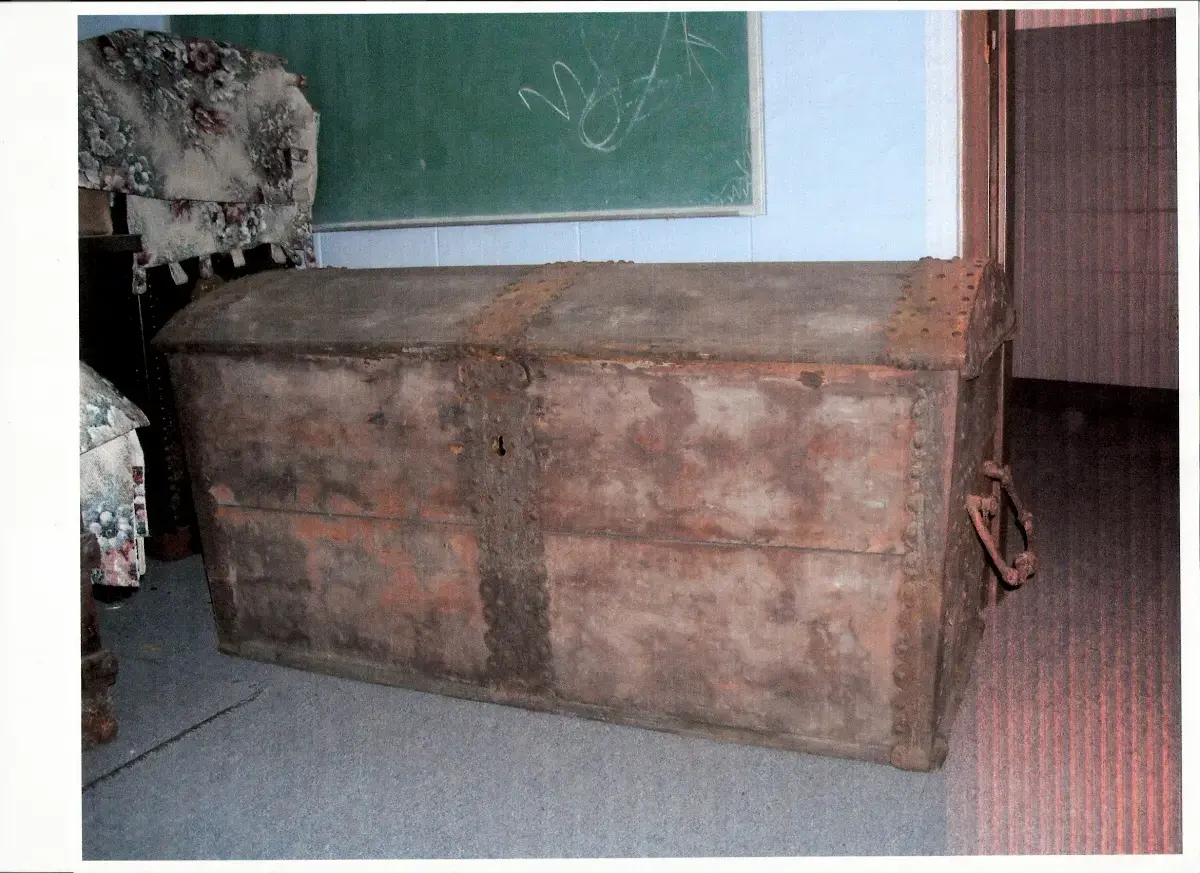 En stor kiste med buet lokk,  jernbeslag og en rester av rødaktig maling står på et gulv. Sammenbrettet, blomstrete tøy til venstre.En grønn tavle på veggen bak. 