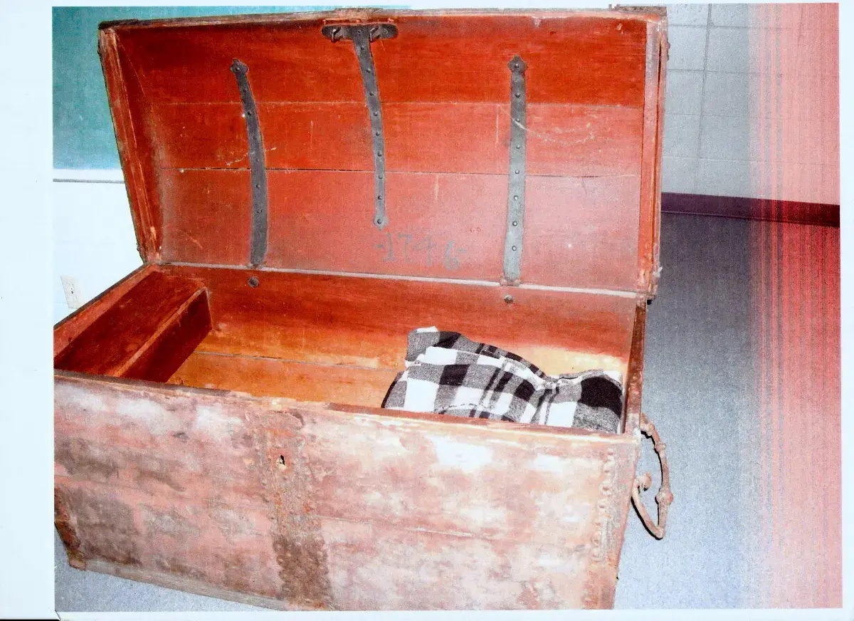 En stor kiste med rødfarger og åpent, buet lokk står på et gulv. Noe sort og hvit rutete ligger i kista.