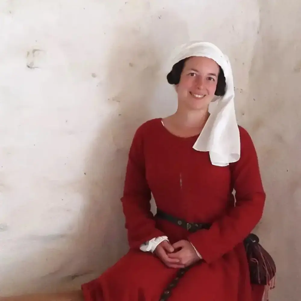 Kvinna i röd medeltida klänning och vit huvudbonad sitter.