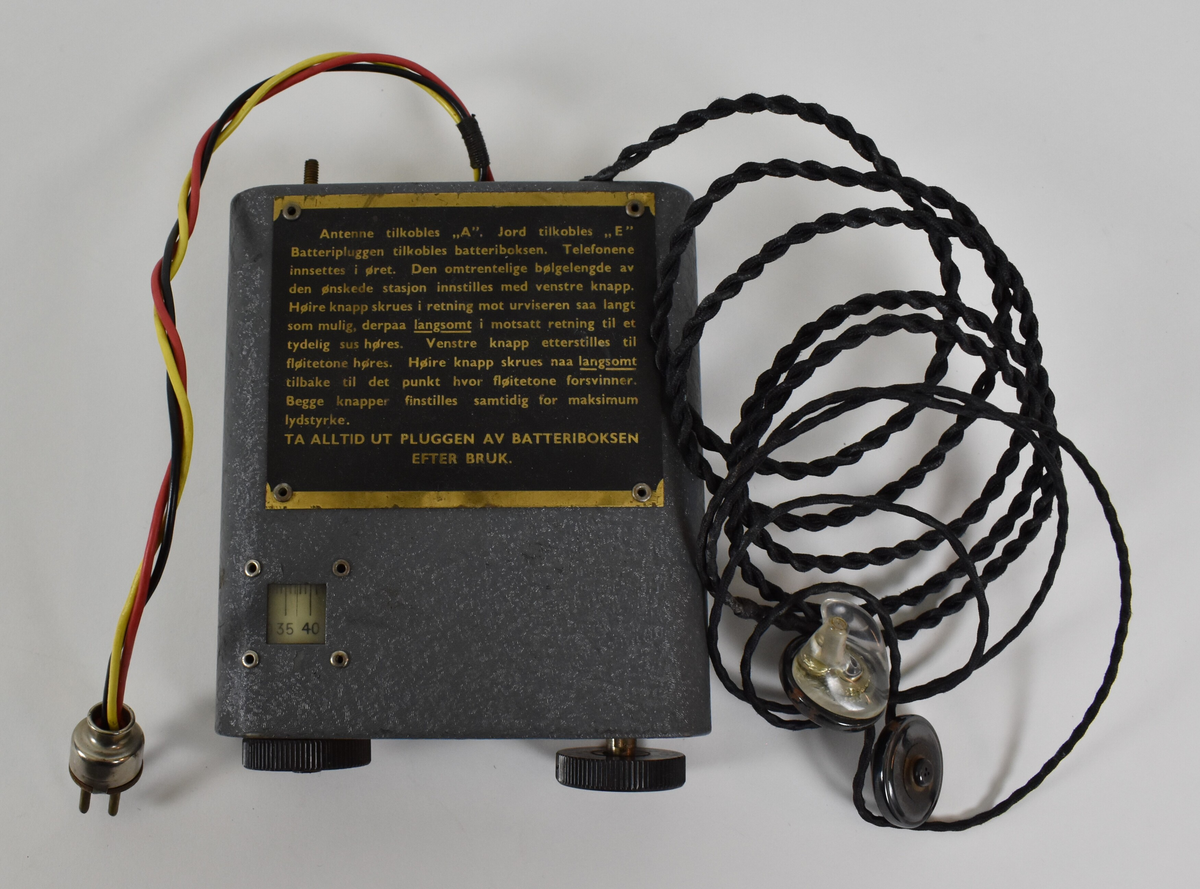 Liten, grå og rektangulær radioboks med to avrundede og to kantede hjørner. Det går to ledninger ut fra den ene enden: den ene ledningen består av en svart, en gul og en rød ledning tvunnet sammen med en stikkontakt i enden, og den andre ledningen består av to svarte ledninger tvunnet sammen med to krystallørepropper i endene. I motsatt ende av boksen er to svarte hjul. Fremsiden av radioen har et lite vindu viser tall til radiostasjoner/bølgelengder. Øverst er et skilt med bruksanvisning for hvordan man kobler radio til batteri og antenne, samt hvordan man bruker radioen for å få lydsignal.