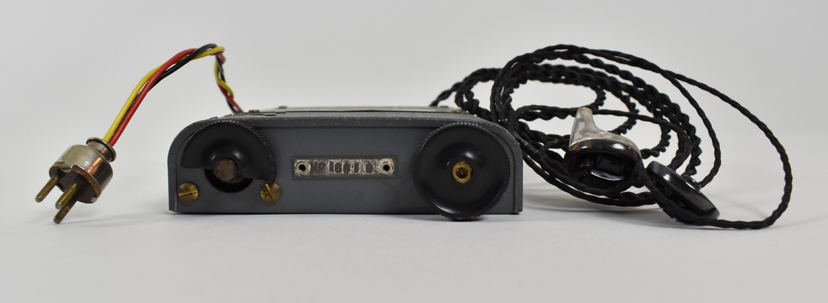 Liten, grå og rektangulær radioboks med to avrundede og to kantede hjørner. Det går to ledninger ut fra den ene enden: den ene ledningen består av en svart, en gul og en rød ledning tvunnet sammen med en stikkontakt i enden, og den andre ledningen består av to svarte ledninger tvunnet sammen med to krystallørepropper i endene. I motsatt ende av boksen er to svarte hjul. Fremsiden av radioen har et lite vindu viser tall til radiostasjoner/bølgelengder. Øverst er et skilt med bruksanvisning for hvordan man kobler radio til batteri og antenne, samt hvordan man bruker radioen for å få lydsignal.