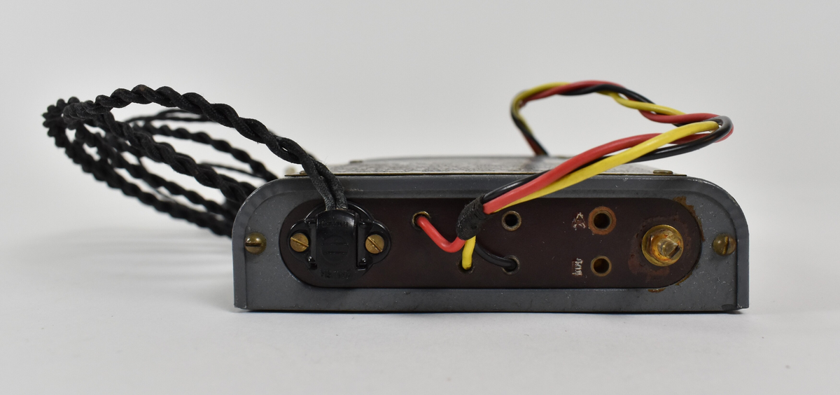 Liten, grå og rektangulær radioboks med to avrundede og to kantede hjørner. Det går to ledninger ut fra den ene enden: den ene ledningen består av en svart, en gul og en rød ledning tvunnet sammen med en stikkontakt i enden, og den andre ledningen består av to svarte ledninger tvunnet sammen med to krystallørepropper i endene. I motsatt ende av boksen er to svarte hjul. Fremsiden av radioen har et lite vindu viser tall til radiostasjoner/bølgelengder. Øverst er et skilt med bruksanvisning for hvordan man kobler radio til batteri og antenne, samt hvordan man bruker radioen for å få lydsignal.