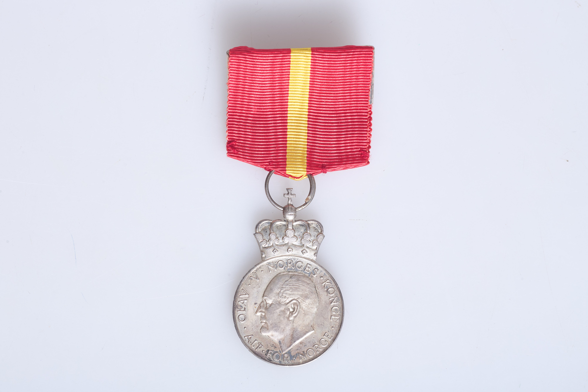 Kongens fortjenestmedalje i sølv (a) med etui (b). Medaljen er festet i et rødt og gult bånd. Tildelt Ludvig Skjerven i 1960. Etuiet dekorert med krone på lokket.