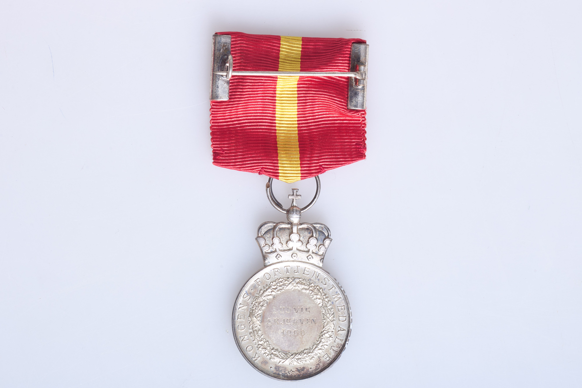 Kongens fortjenestmedalje i sølv (a) med etui (b). Medaljen er festet i et rødt og gult bånd. Tildelt Ludvig Skjerven i 1960. Etuiet dekorert med krone på lokket.