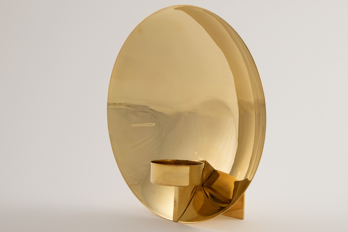 Vegglampe i messing. Rund parabolformet skive med telysholder foran. Rektangulært feste på baksiden med en avstandsplate mellom fest og parabol. Designet tar opp i seg den pre-elektriske lampetypen med reflektorskjerm.