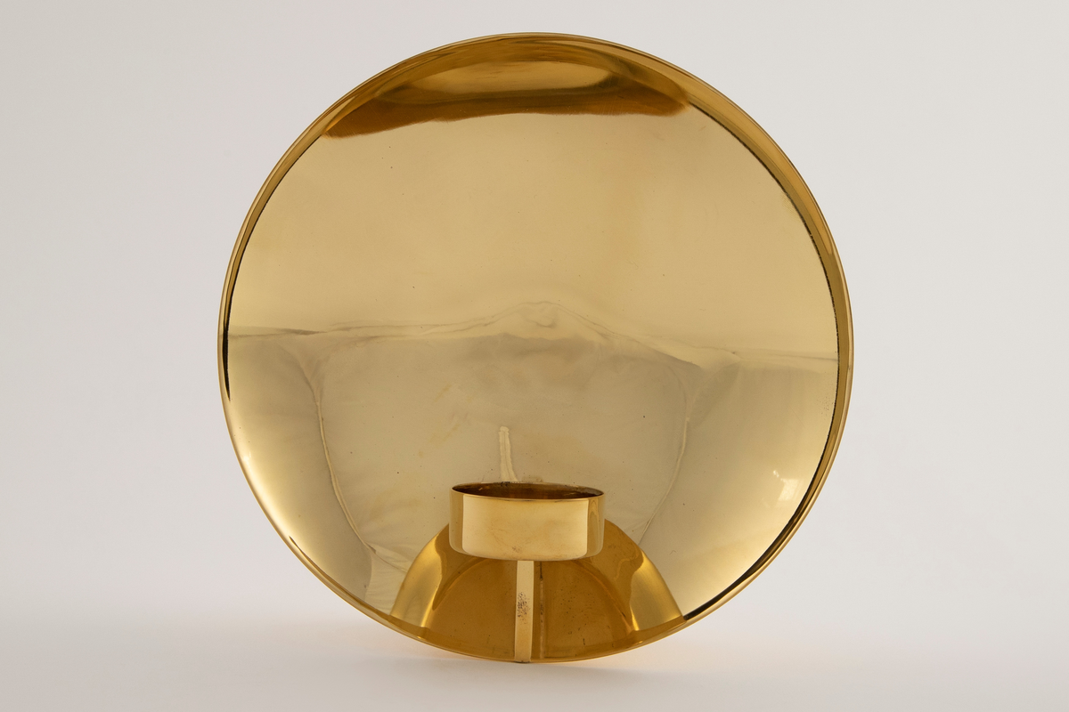 Vegglampe i messing. Rund parabolformet skive med telysholder foran. Rektangulært feste på baksiden med en avstandsplate mellom fest og parabol. Designet tar opp i seg den pre-elektriske lampetypen med reflektorskjerm.