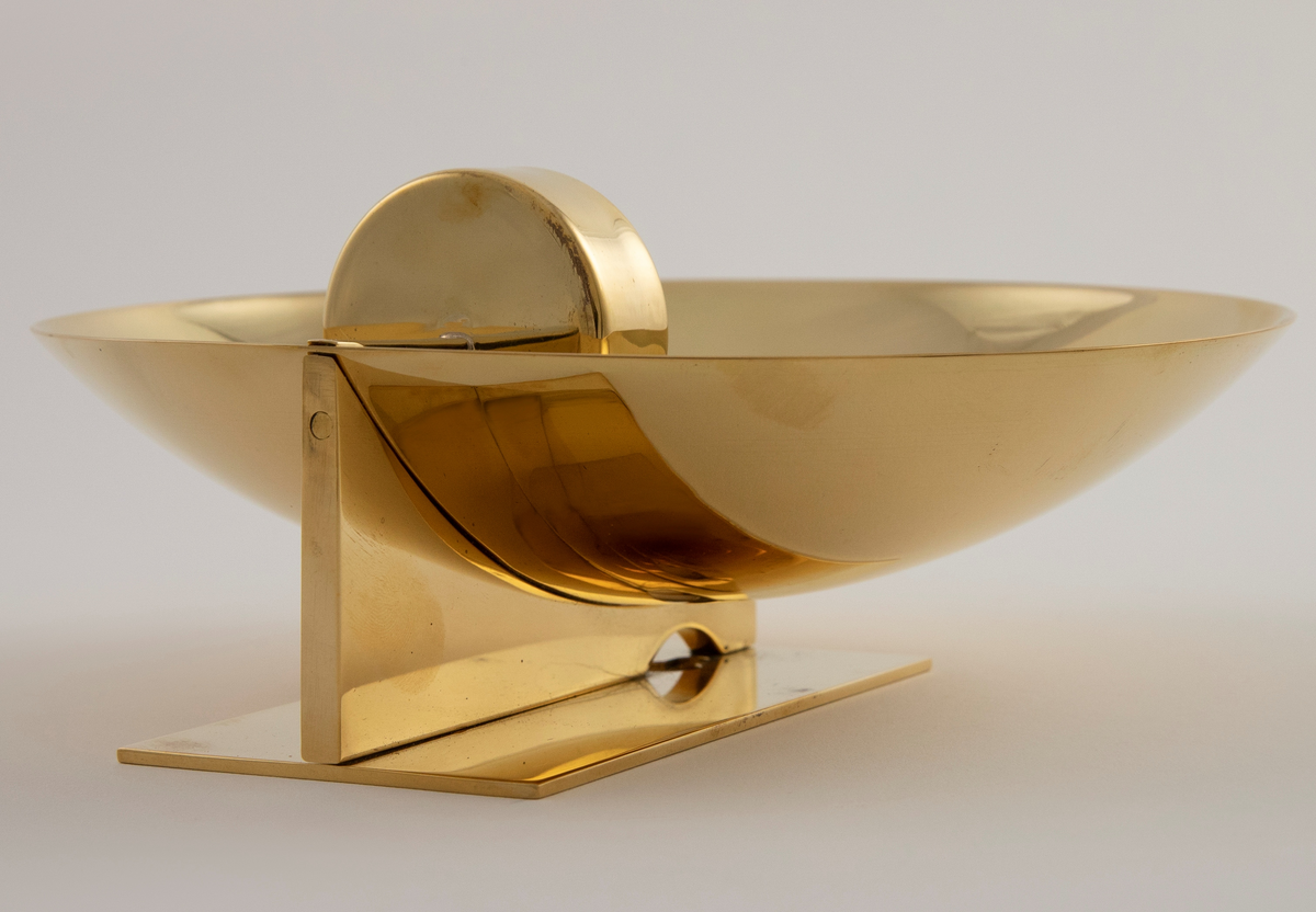 Vegglampe i messing. Rund parabolformet skive med telysholder foran. Rektangulært feste på baksiden med en avstandsplate mellom fest og parabol. Designet tar opp i seg den pre-elektriske lampetypen med reflektorskjerm.