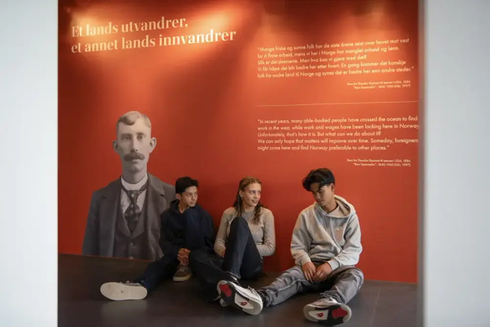 Tre ungdommer sitter foran en oransje utstillingsvegg med tittel Et lands utvandrer er et annet lands innvandrer.