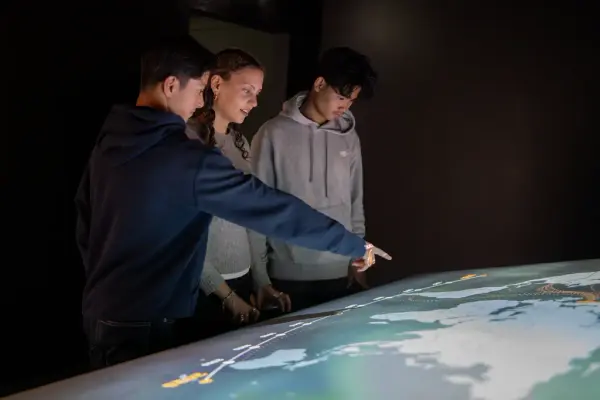 Tre ungdommer ser på et opplyst verdenskart. Gutten til venstre peker på kartet.