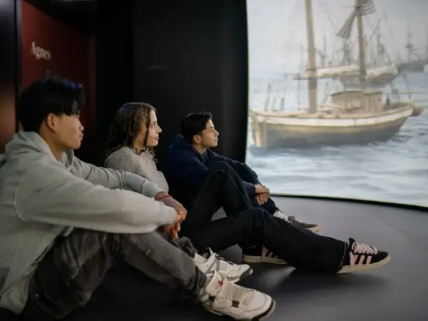 Tre ungdommer sitter på gulvet til venstre. Til høyre en skjerm med bilde av flere seiskuter.