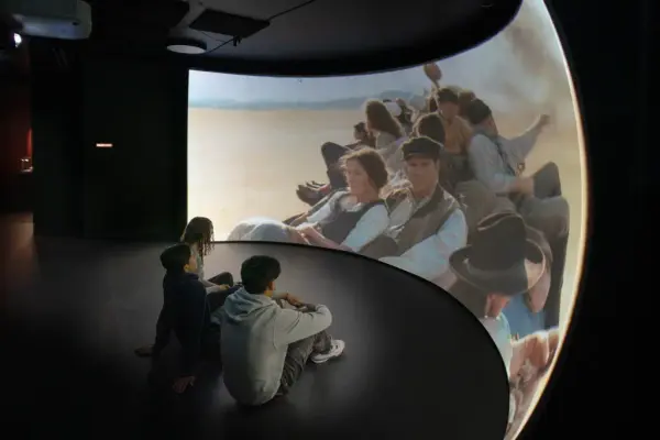 Tre ungdommer sitter på gulvet foran en buet skjerm der et filmbilde vises av en gruppe mennesker.