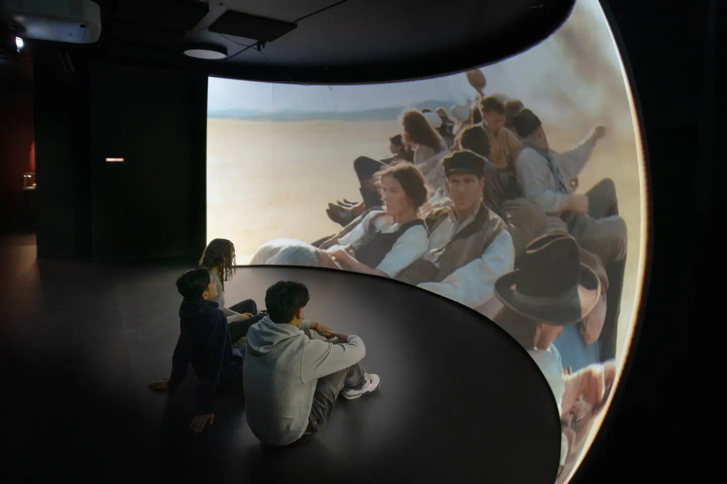 Tre ungdommer sitter på gulvet foran en buet skjerm der et filmbilde vises av en gruppe mennesker.