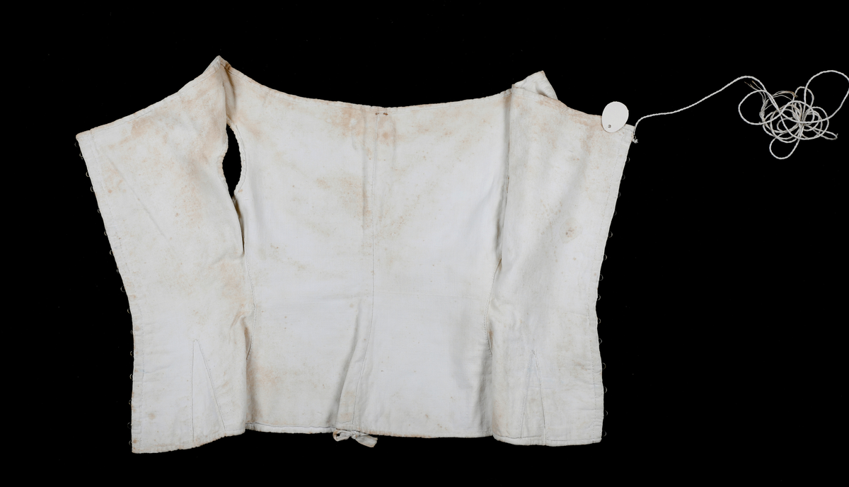 Liv eller korsett av lin med metallhekter og tvunnet tråd til snøring. Det har søm midt bak med hempe oppe og nede. Sidesømmen er litt tilbaketrukket, og det er felt inn stoff neders i sida for utsving fra livet. Halsåpningen er båtformet.