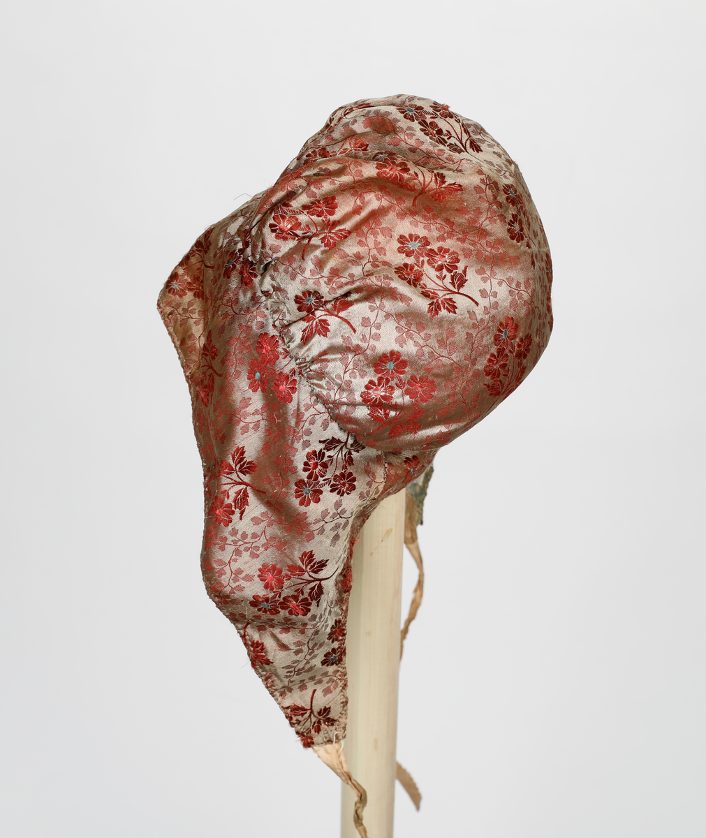 Høglue i mønstret silke med beige knytebånd som er sikk-sakk-klippet i kantene. Lua er fòret med smårutete bomullsstoff.  Pannestykket er sydd i to hoveddeler der det er skjøtt inn to stykker i den ene halvdelen. Rundt kanten er det sydd kastesting med prikk. Pullen er rynket til pannestykket.