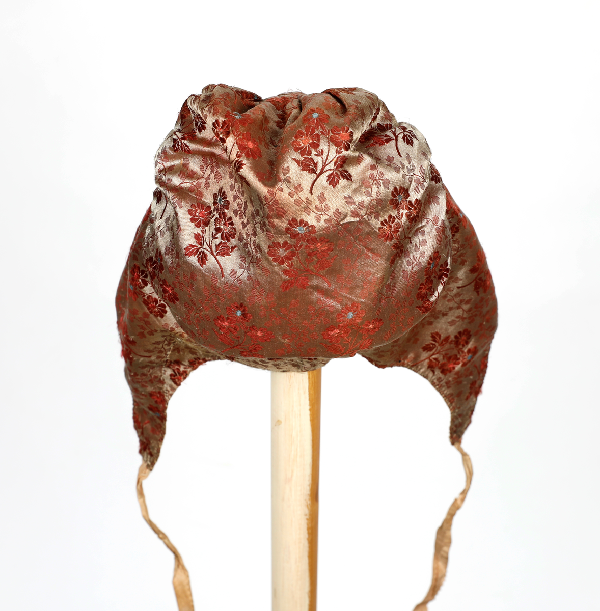 Høglue i mønstret silke med beige knytebånd som er sikk-sakk-klippet i kantene. Lua er fòret med smårutete bomullsstoff.  Pannestykket er sydd i to hoveddeler der det er skjøtt inn to stykker i den ene halvdelen. Rundt kanten er det sydd kastesting med prikk. Pullen er rynket til pannestykket.