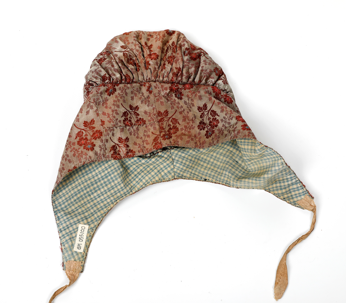 Høglue i mønstret silke med beige knytebånd som er sikk-sakk-klippet i kantene. Lua er fòret med smårutete bomullsstoff.  Pannestykket er sydd i to hoveddeler der det er skjøtt inn to stykker i den ene halvdelen. Rundt kanten er det sydd kastesting med prikk. Pullen er rynket til pannestykket.