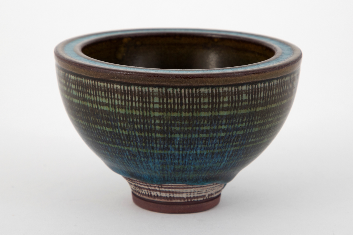 Rund skål i steingods. Konkav form med tykke vegger og lavt fotstykke. På utsiden er radiære linjer risset inn i godset og overflaten dekket med horisontale hvite ringer. Skålen er dekket med glasur i grønne og turkise nyanser med en krakelert virkning. Den turkise fargen er konsentrert om bunnen innvendig, munningskanten samt fotstykket utvendig. På fotstykket har glasuren rent ujevnt og et stykke av godset ligger blottet.