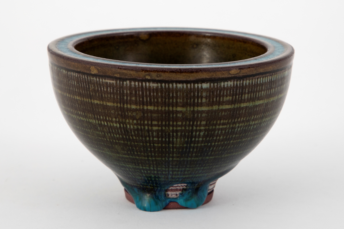 Rund skål i steingods. Konkav form med tykke vegger og lavt fotstykke. På utsiden er radiære linjer risset inn i godset og overflaten dekket med horisontale hvite ringer. Skålen er dekket med glasur i grønne og turkise nyanser med en krakelert virkning. Den turkise fargen er konsentrert om bunnen innvendig, munningskanten samt fotstykket utvendig. På fotstykket har glasuren rent ujevnt og et stykke av godset ligger blottet.