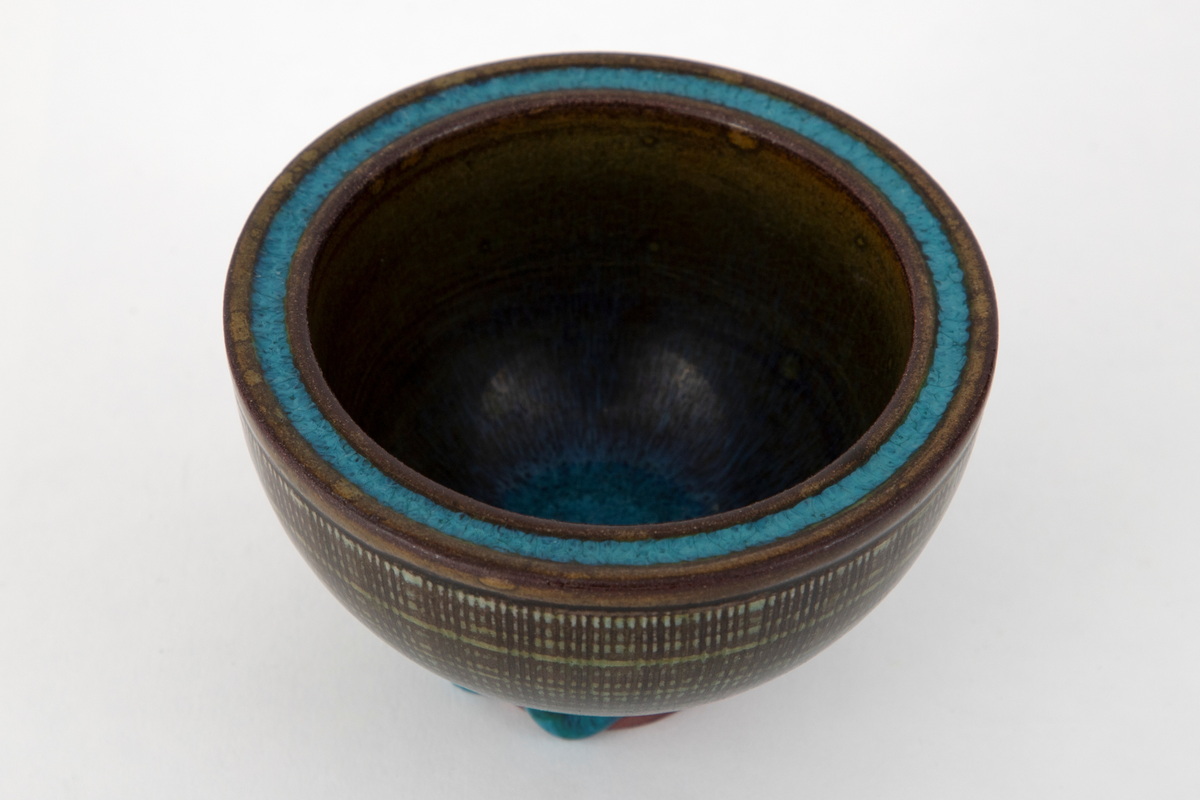 Rund skål i steingods. Konkav form med tykke vegger og lavt fotstykke. På utsiden er radiære linjer risset inn i godset og overflaten dekket med horisontale hvite ringer. Skålen er dekket med glasur i grønne og turkise nyanser med en krakelert virkning. Den turkise fargen er konsentrert om bunnen innvendig, munningskanten samt fotstykket utvendig. På fotstykket har glasuren rent ujevnt og et stykke av godset ligger blottet.