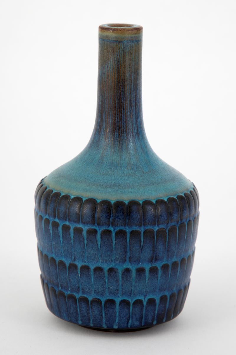 Flaskeformet krukke i steingods, Tilnærmet sylinderformet korpus, svakt utvidet oppad. Kraftig innsnevring ved overgang til halspartiet som har en smal sylinderform. Korpus er dekorert med regelmessige horisontale rekker med ovale former. Vasen er dekket med en glasur i blå nyanser, noe brun på halsen og sort på deler av de ovale formene.