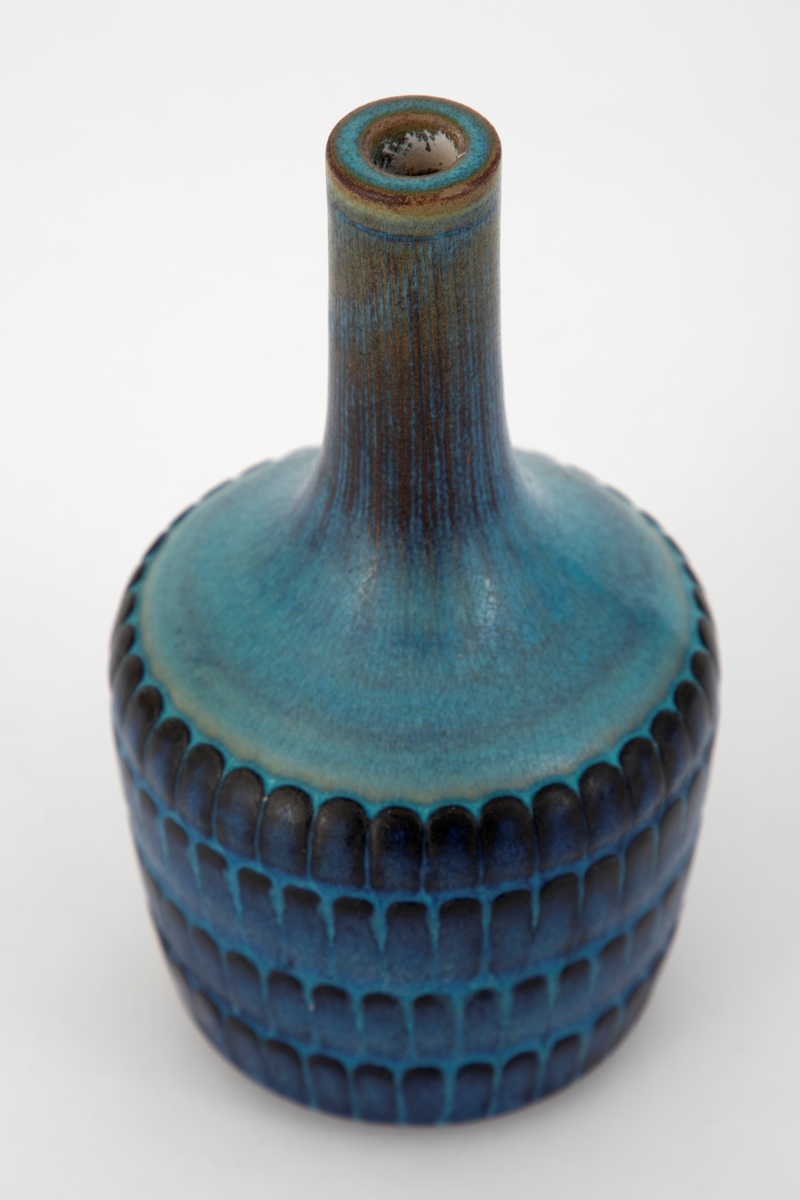 Flaskeformet krukke i steingods, Tilnærmet sylinderformet korpus, svakt utvidet oppad. Kraftig innsnevring ved overgang til halspartiet som har en smal sylinderform. Korpus er dekorert med regelmessige horisontale rekker med ovale former. Vasen er dekket med en glasur i blå nyanser, noe brun på halsen og sort på deler av de ovale formene.