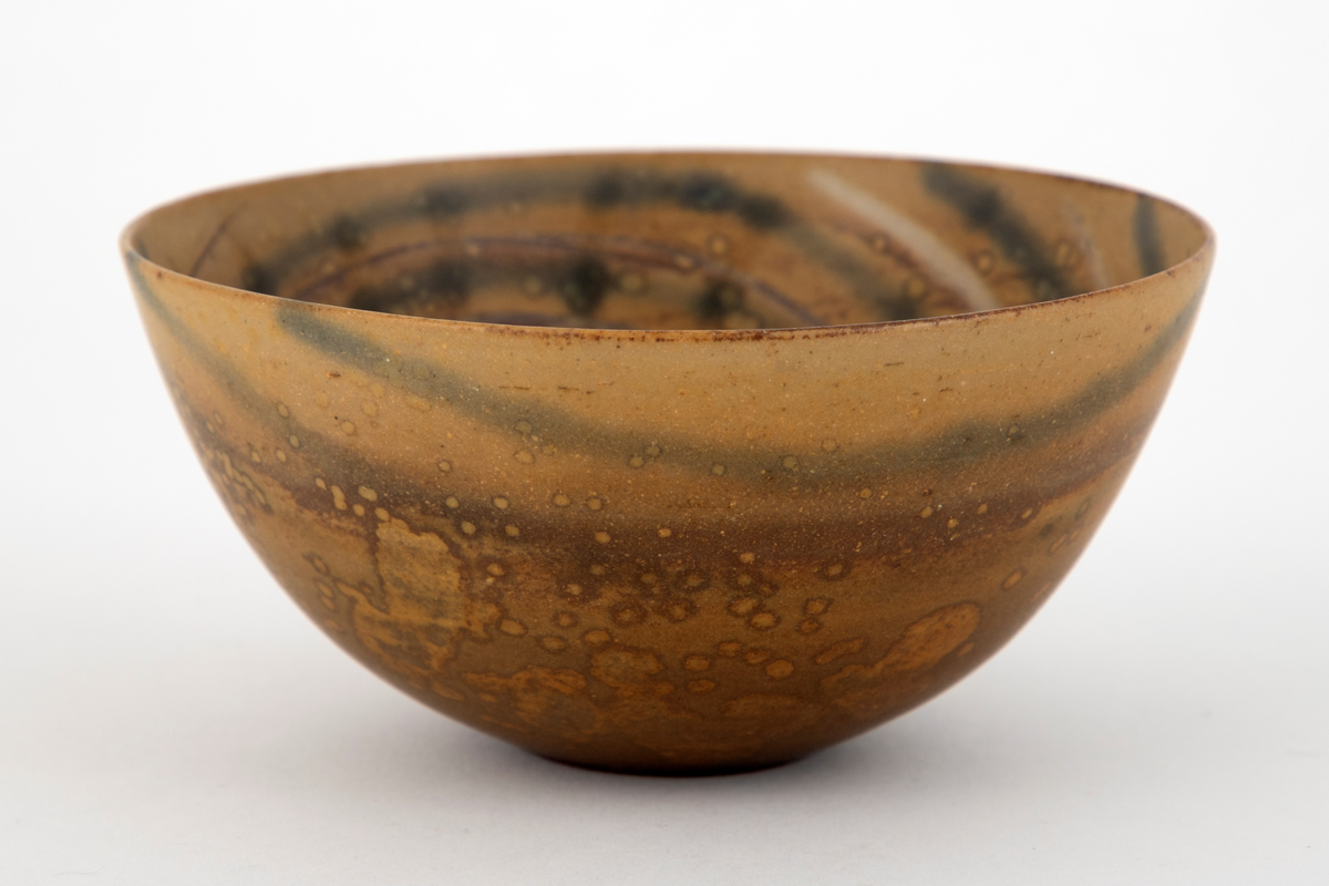 Skål i steingods. Konkav form med flat bunn. Skålen er dekket med brunspettet, okerfarge glasur. Innvendig et lysere mønster av konsentriske sirkler, forskjøvet mot den ene siden.