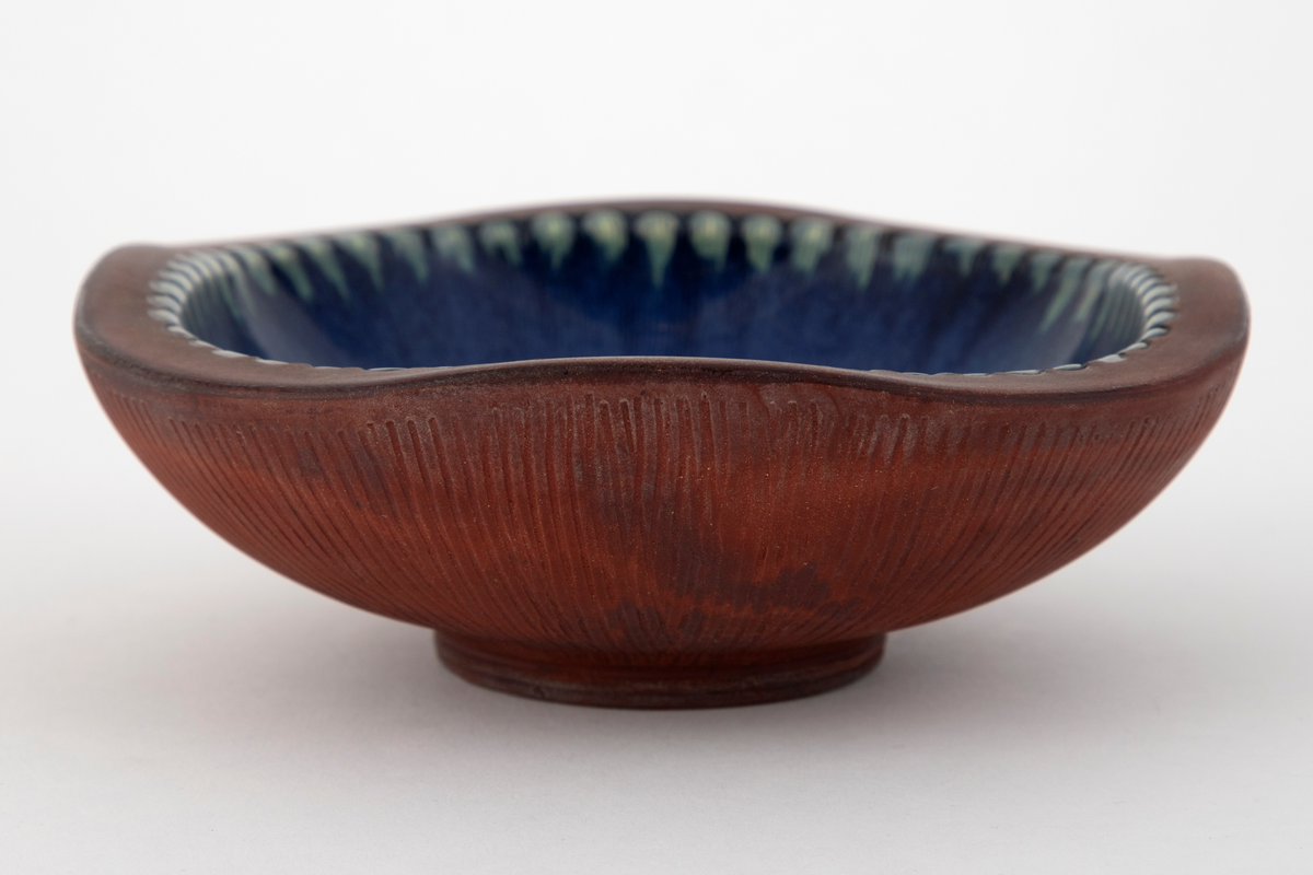 Skål i steingods. Skålens bunnform er rund, men på grunn av flattrykte sider er skålen nærmest firkantet ved munningsranden. Utvendig er skålen uglasert, men dekorert med innrissede, radiære rifler utgående fra fotringen. Den tykke munningsranden er også uglasert. Innvendig er skålen glasert med blå glasur, med en brun flekk i bunnen. Langs randen en rekke med tungeformede fordypninger glasert i lysegrønt.