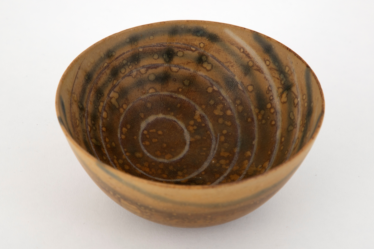 Skål i steingods. Konkav form med flat bunn. Skålen er dekket med brunspettet, okerfarge glasur. Innvendig et lysere mønster av konsentriske sirkler, forskjøvet mot den ene siden.