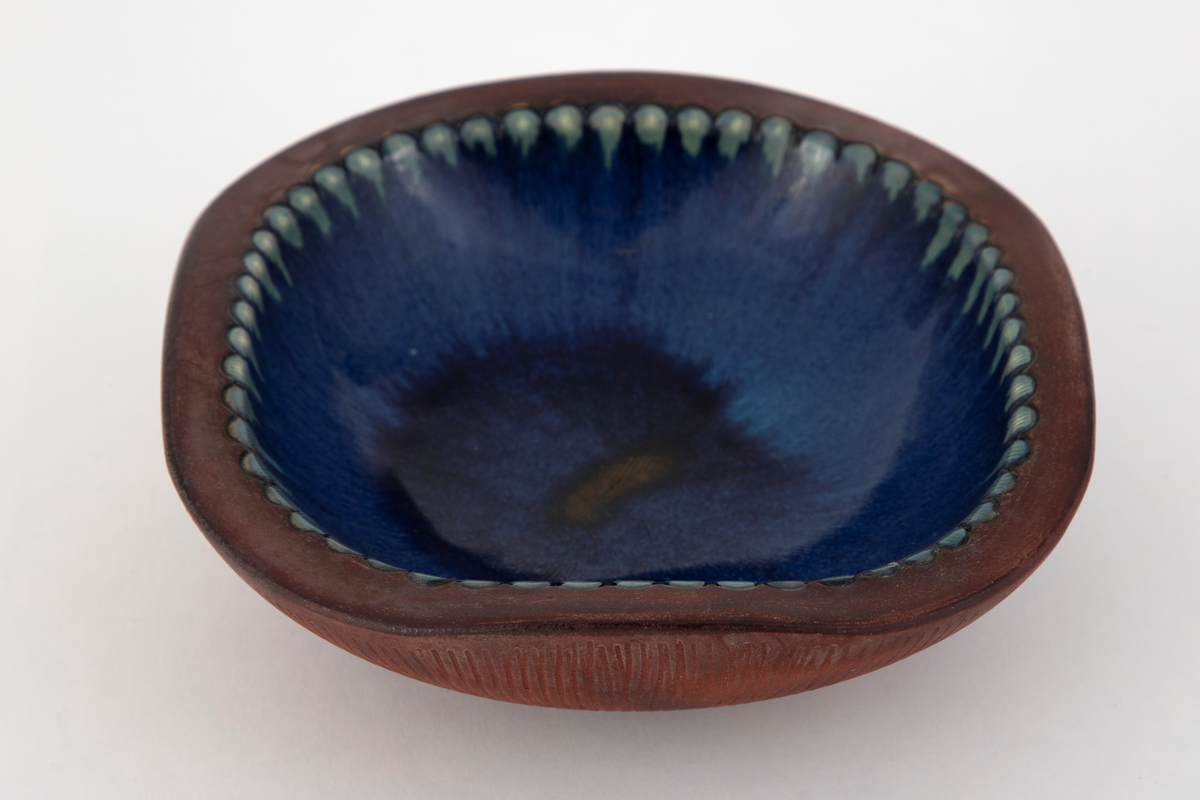 Skål i steingods. Skålens bunnform er rund, men på grunn av flattrykte sider er skålen nærmest firkantet ved munningsranden. Utvendig er skålen uglasert, men dekorert med innrissede, radiære rifler utgående fra fotringen. Den tykke munningsranden er også uglasert. Innvendig er skålen glasert med blå glasur, med en brun flekk i bunnen. Langs randen en rekke med tungeformede fordypninger glasert i lysegrønt.