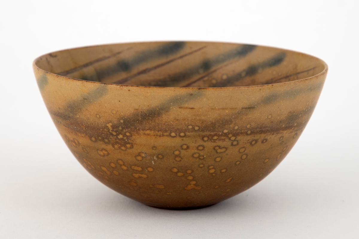 Skål i steingods. Konkav form med flat bunn. Skålen er dekket med brunspettet, okerfarge glasur. Innvendig et lysere mønster av konsentriske sirkler, forskjøvet mot den ene siden.