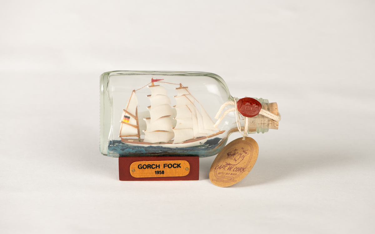 Flaskeskute med en hvit skute inni. Skipet har tysk flagg og alle seil oppe. Under skuta er det blå bølger. Flaskeskuta har pålimt sokkel i tre med navneplakett på. I flasketuten er det en kork og et rødt vokssegl. Der henger det også en merkelapp med garantiinformasjon.