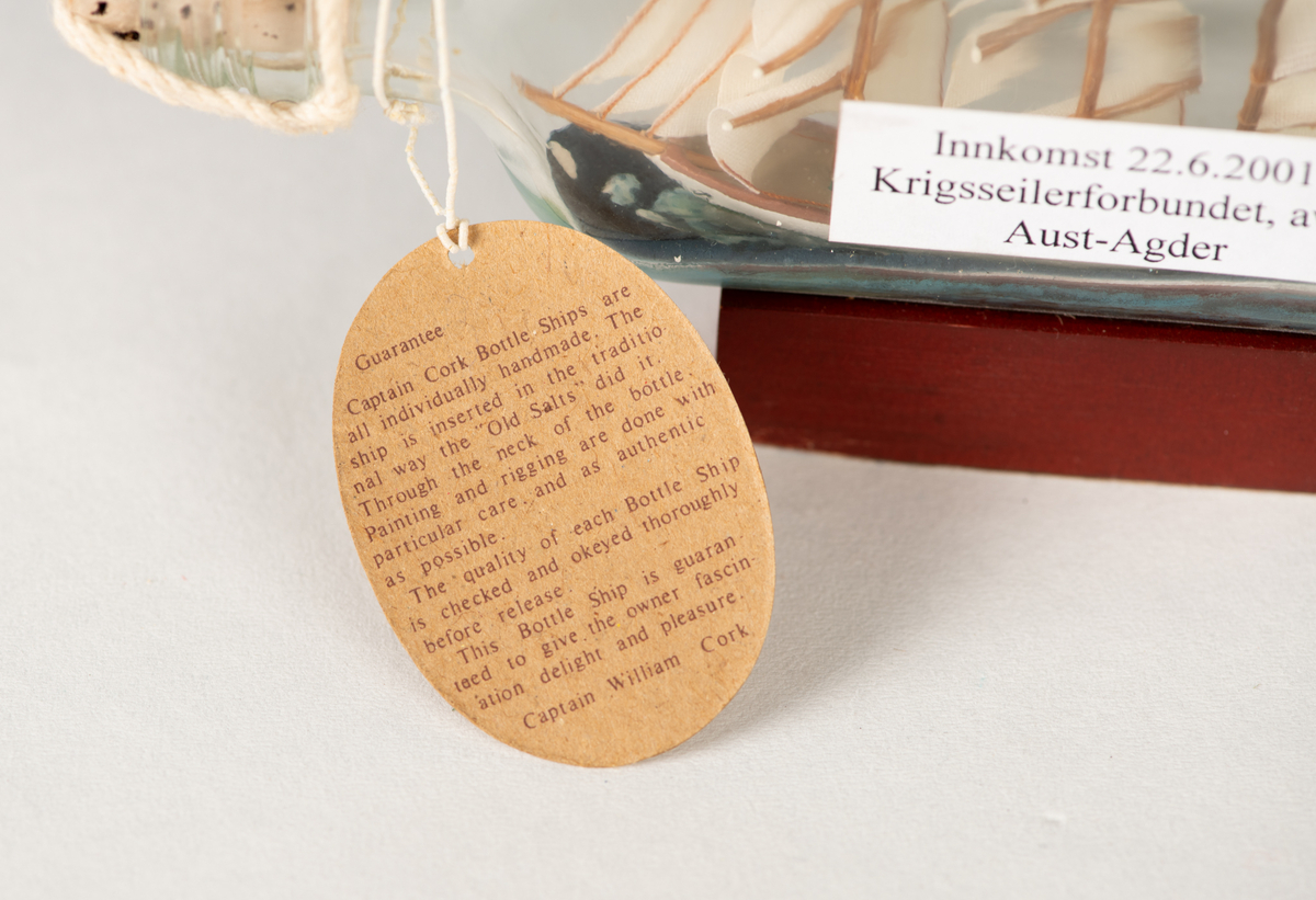 Flaskeskute med en hvit skute inni. Skipet har tysk flagg og alle seil oppe. Under skuta er det blå bølger. Flaskeskuta har pålimt sokkel i tre med navneplakett på. I flasketuten er det en kork og et rødt vokssegl. Der henger det også en merkelapp med garantiinformasjon.