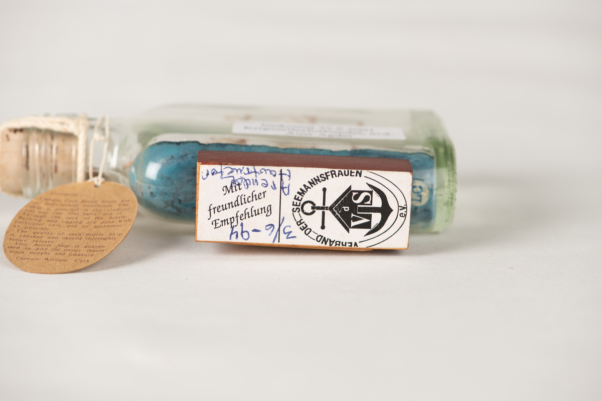 Flaskeskute med en hvit skute inni. Skipet har tysk flagg og alle seil oppe. Under skuta er det blå bølger. Flaskeskuta har pålimt sokkel i tre med navneplakett på. I flasketuten er det en kork og et rødt vokssegl. Der henger det også en merkelapp med garantiinformasjon.