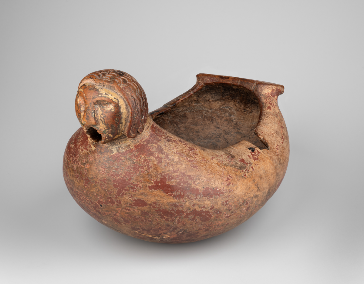 Trøys av bjørk. Oval form. Utskåret mannshode med munnåpning. Håndtak med hakkformete utskjæringer og innskrift. Svarte bekklumper i bunnen.