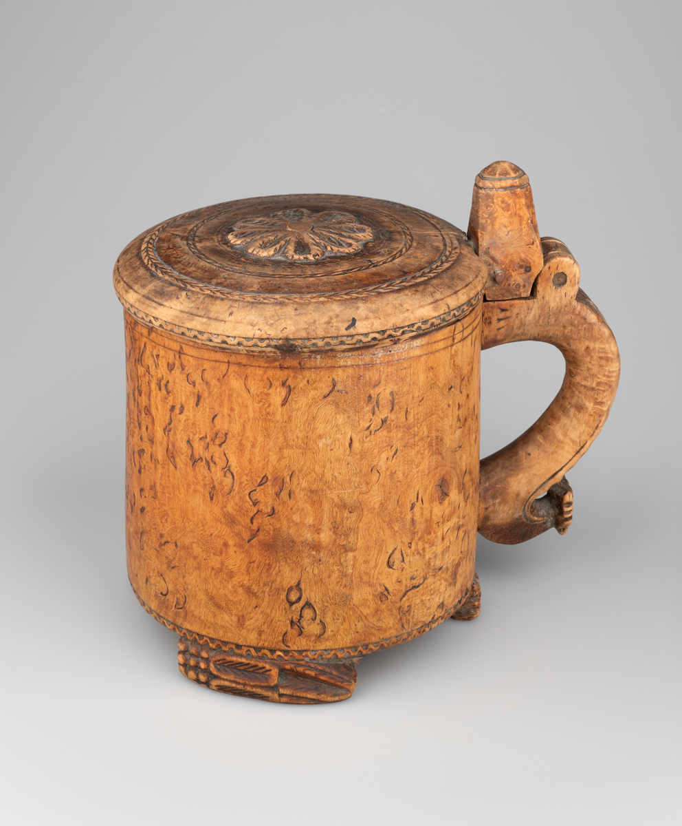 Kanne av dreid valbjørk. Lokk med skjært rosett og to bårder. Tre føtter med utskjæringer.