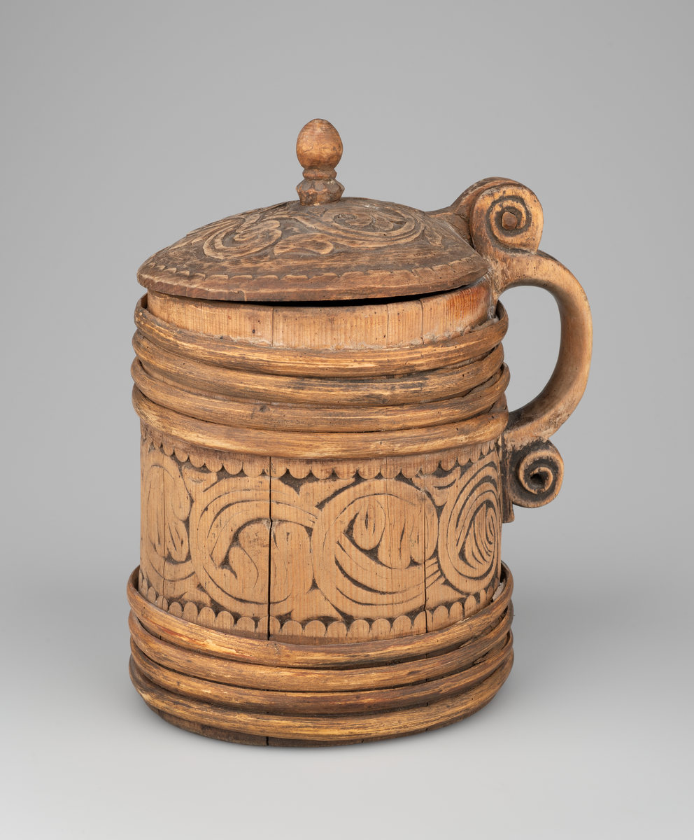 Kanne av trestaver. Lokket med bladlignende ranke, tungeformet bård langs kanten. Lokkgrepet er øverst kuleformet, nederst en ring  med skulpelignende utskjæringer. Hank med spiralformede utskjæringer oppe og nede, er i ett med en av stavene. Korpus har fire gjorder oppe og nede, mellom gjordene en akantusranke, like under og over gjordene tungeformede utskjæringer.