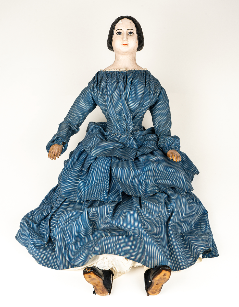 Docka, "Fedora", kroppen är svarvad av prosten Carl Steffenburg, Sundborn (född 1799), är iklädd linne, snörliv, byxor, stupp, tre underkjolar och blå klänning med vita prickar.
Kjolen har två volanger, skörtet på livstycket bildar en tredje volang.