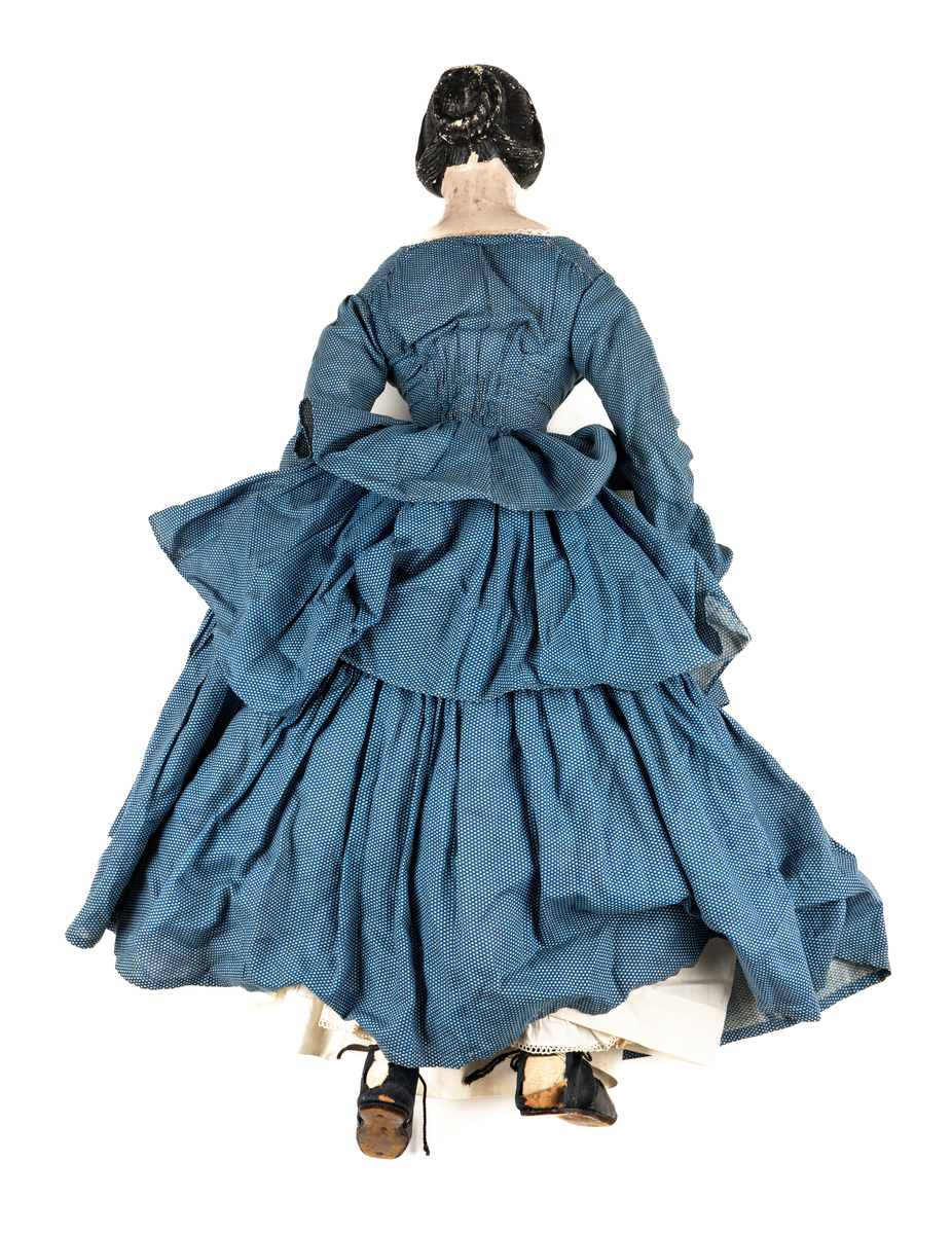 Docka, "Fedora", kroppen är svarvad av prosten Carl Steffenburg, Sundborn (född 1799), är iklädd linne, snörliv, byxor, stupp, tre underkjolar och blå klänning med vita prickar.
Kjolen har två volanger, skörtet på livstycket bildar en tredje volang.