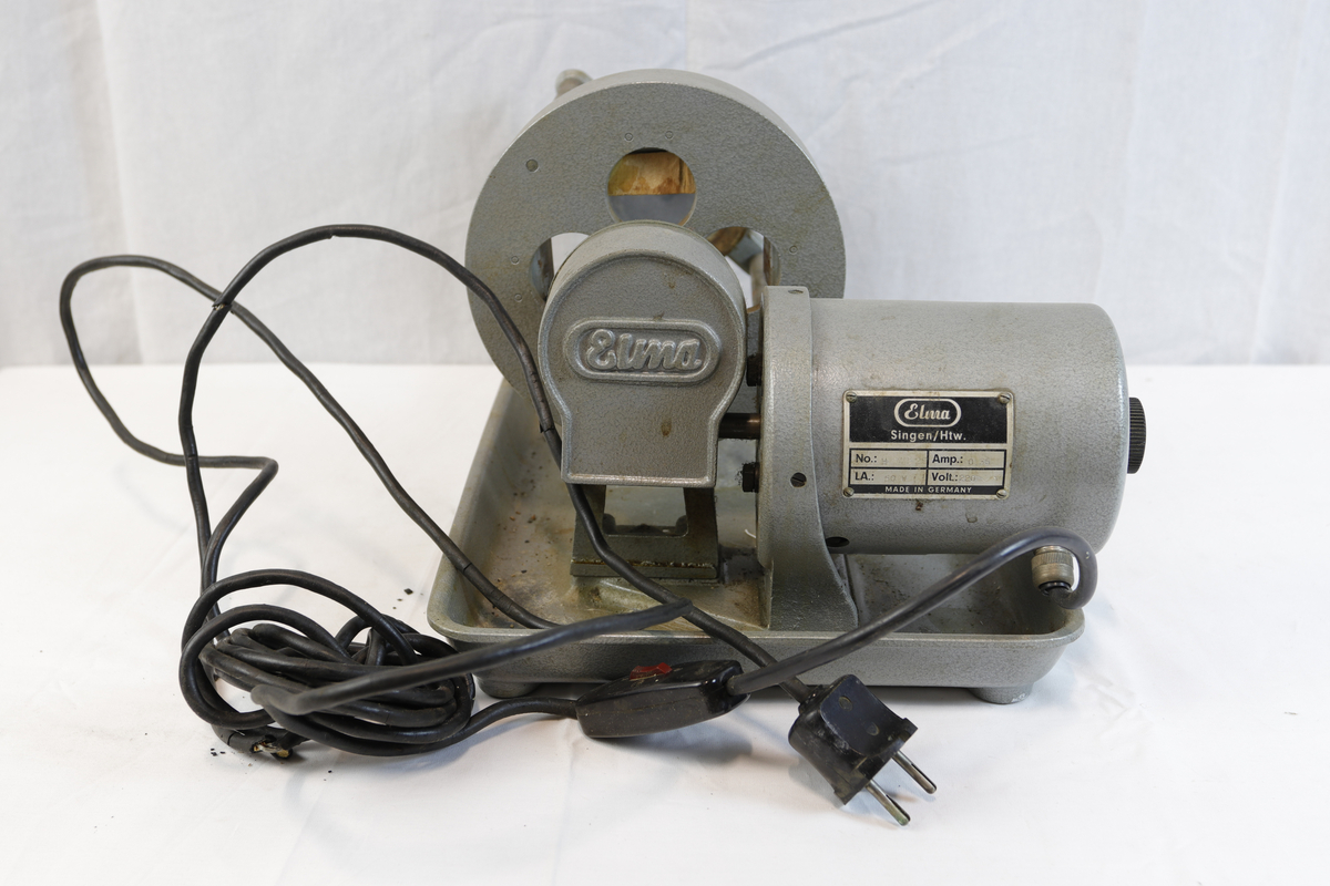 Slipemaskin fra merket Elma. Maskninen er av eldre standard og har stikkontakt med rød bryter. Slipemaskinen har motor og beltedrift, montert på metallbrett. Har roterende hjul og justerbar arm for sliping eller polering av materialer.