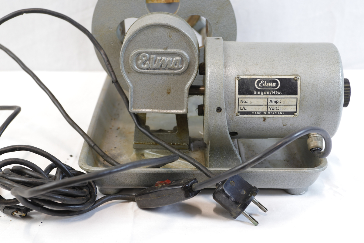 Slipemaskin fra merket Elma. Maskninen er av eldre standard og har stikkontakt med rød bryter. Slipemaskinen har motor og beltedrift, montert på metallbrett. Har roterende hjul og justerbar arm for sliping eller polering av materialer.