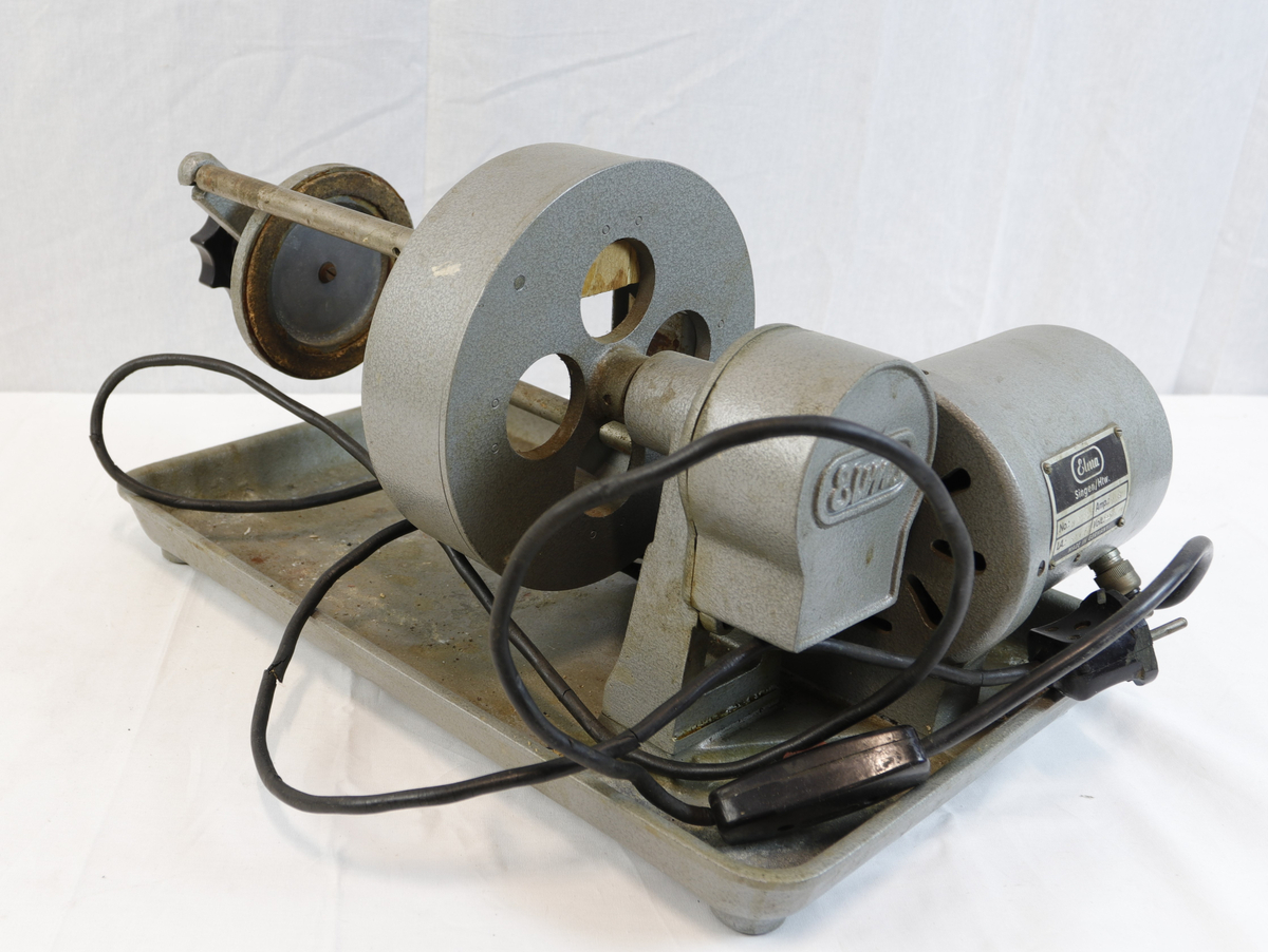 Slipemaskin fra merket Elma. Maskninen er av eldre standard og har stikkontakt med rød bryter. Slipemaskinen har motor og beltedrift, montert på metallbrett. Har roterende hjul og justerbar arm for sliping eller polering av materialer.