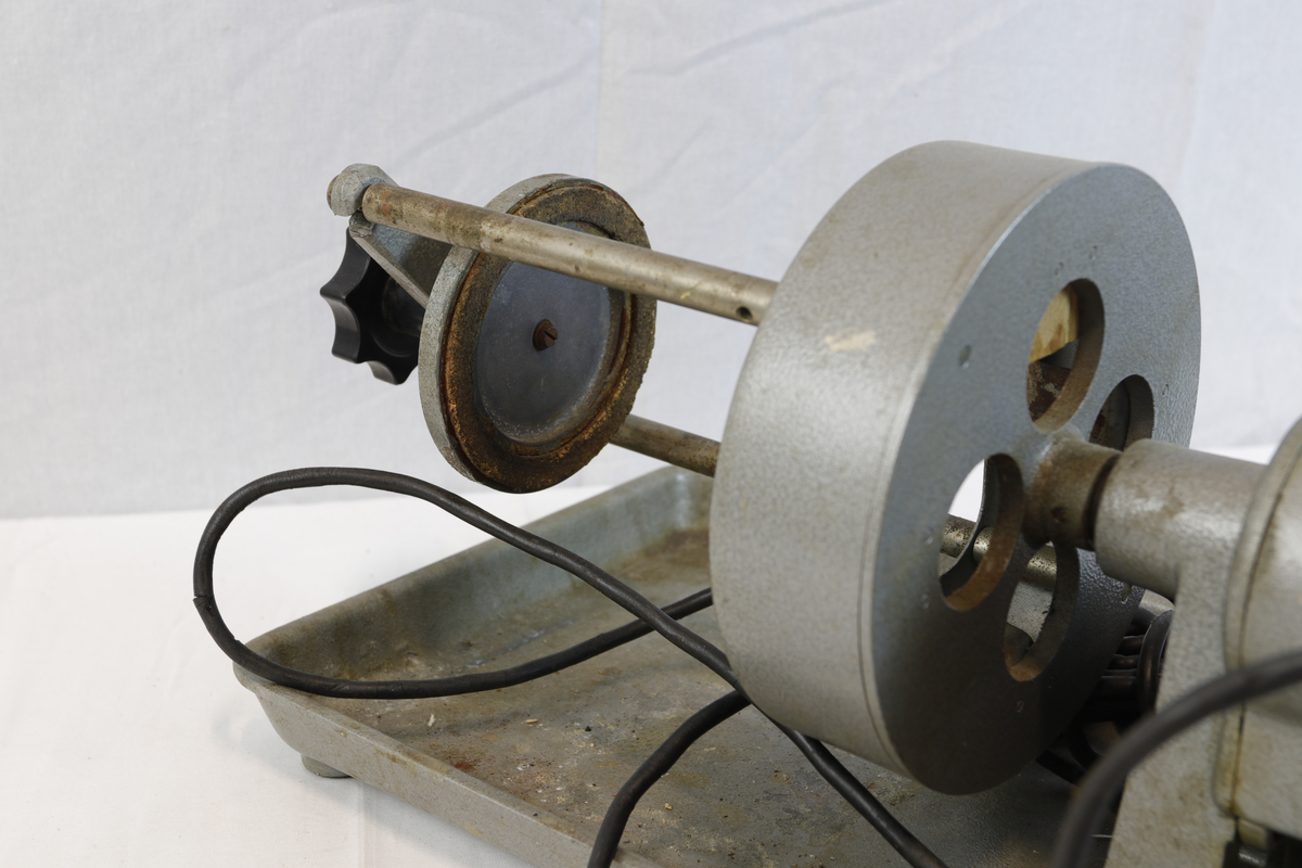 Slipemaskin fra merket Elma. Maskninen er av eldre standard og har stikkontakt med rød bryter. Slipemaskinen har motor og beltedrift, montert på metallbrett. Har roterende hjul og justerbar arm for sliping eller polering av materialer.