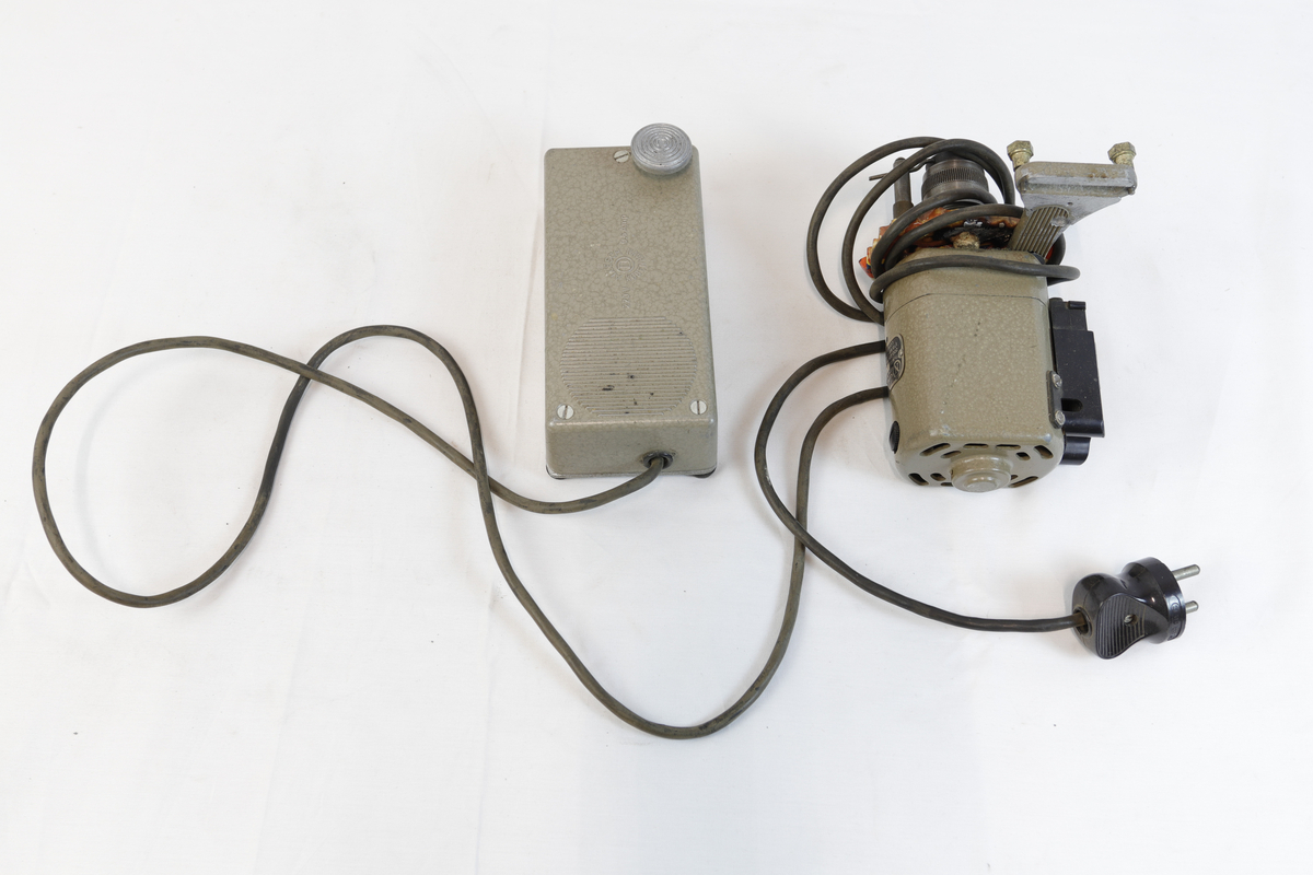 Elektrisk enhet med kontrollboks og motor, koblet sammen med ledninger. Motoren har sylindrisk form og er festet til en brakett – trolig del av et eldre verktøy eller maskin. Den har et skilt, som betegner den som "symotor". 