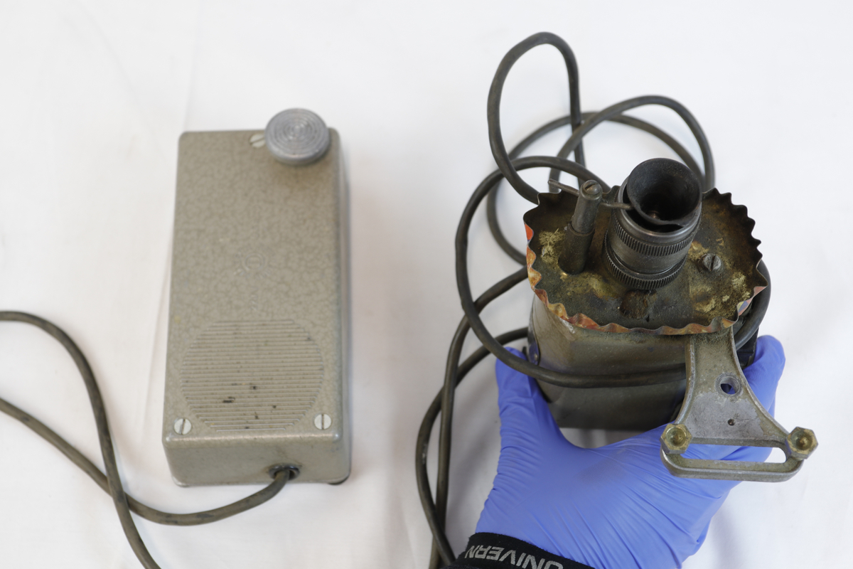 Elektrisk enhet med kontrollboks og motor, koblet sammen med ledninger. Motoren har sylindrisk form og er festet til en brakett – trolig del av et eldre verktøy eller maskin. Den har et skilt, som betegner den som "symotor". 