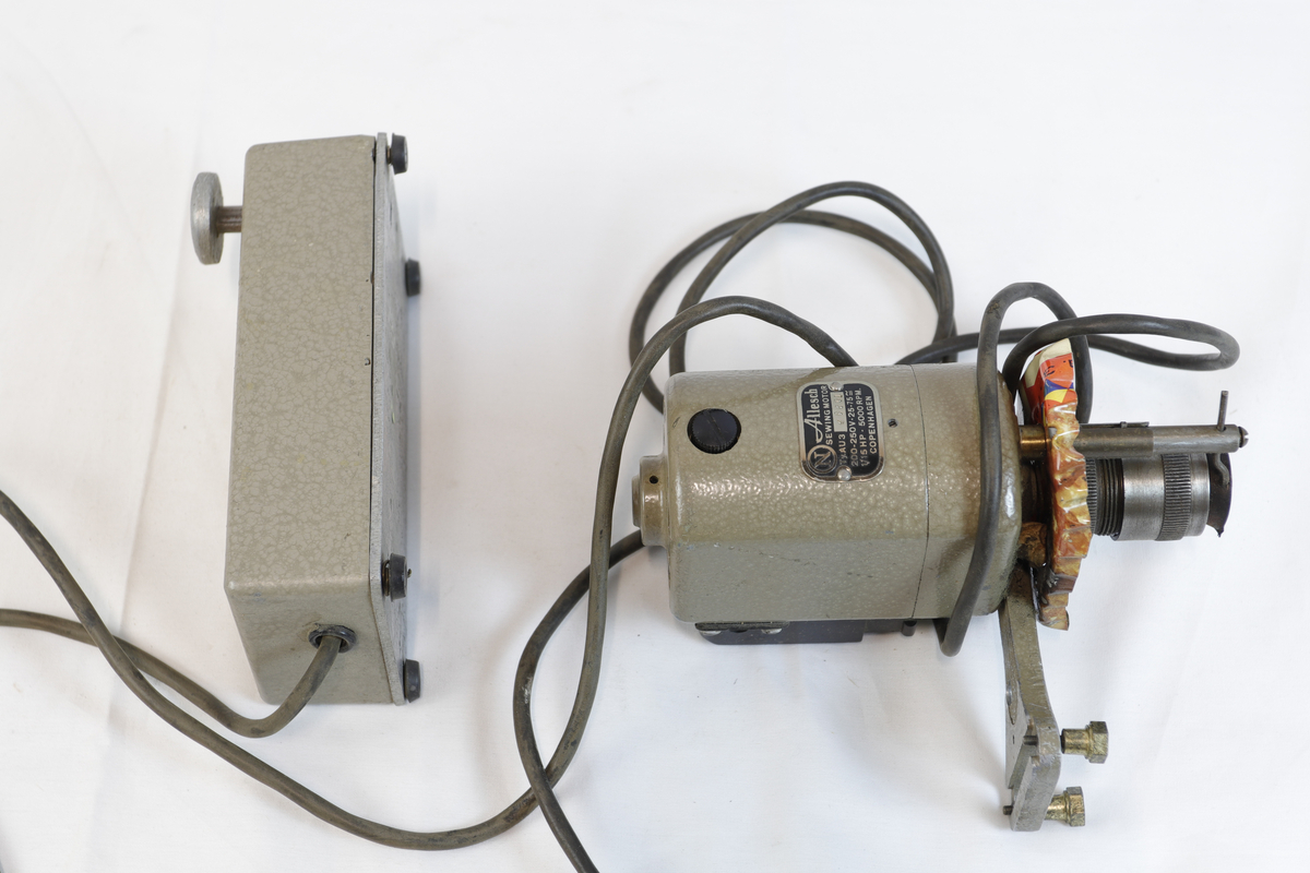 Elektrisk enhet med kontrollboks og motor, koblet sammen med ledninger. Motoren har sylindrisk form og er festet til en brakett – trolig del av et eldre verktøy eller maskin. Den har et skilt, som betegner den som "symotor". 