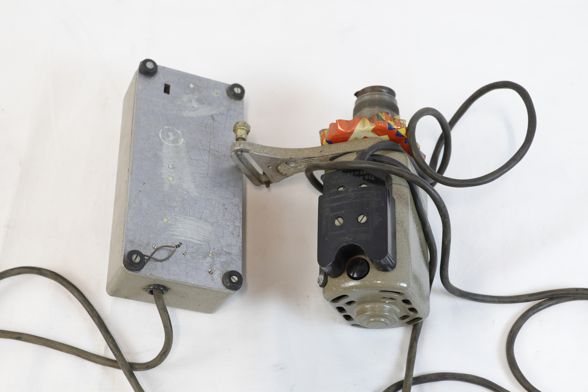 Elektrisk enhet med kontrollboks og motor, koblet sammen med ledninger. Motoren har sylindrisk form og er festet til en brakett – trolig del av et eldre verktøy eller maskin. Den har et skilt, som betegner den som "symotor". 