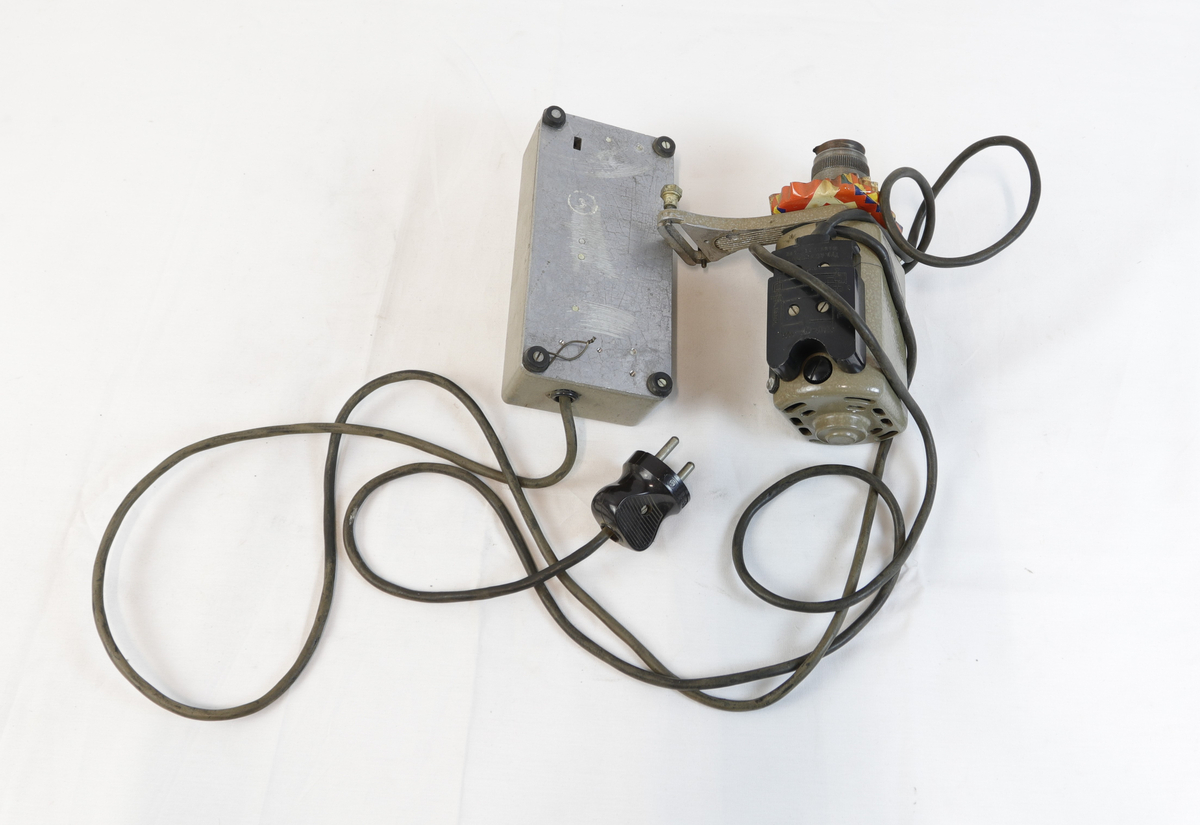 Elektrisk enhet med kontrollboks og motor, koblet sammen med ledninger. Motoren har sylindrisk form og er festet til en brakett – trolig del av et eldre verktøy eller maskin. Den har et skilt, som betegner den som "symotor". 