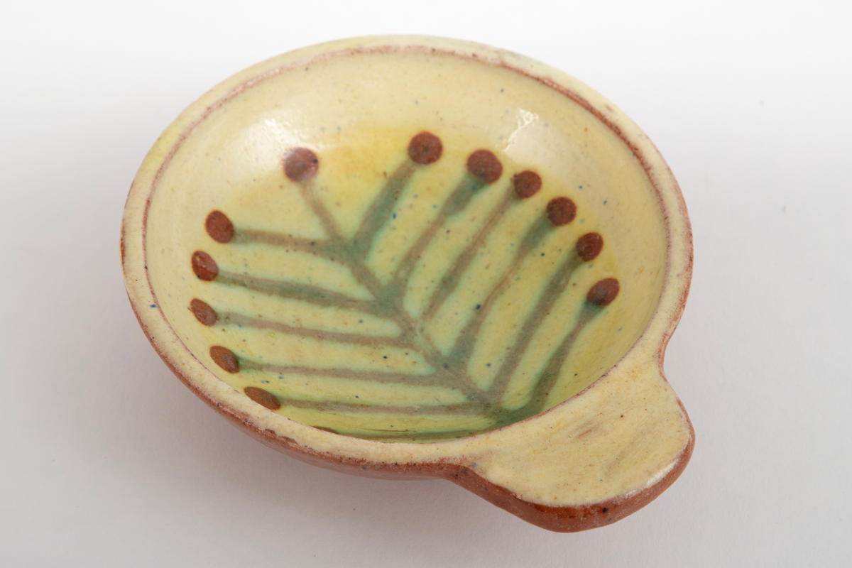 Kabaretskål av dreid leirgods. Skålene er runde med en enkel flat hank. På innsiden har skålen gulhvit glasur og er dekorert med en grønne gren.
